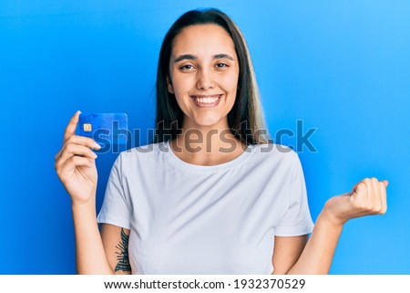 Young hispanic woman holding credit card screaming proud, celebrating victory and success very excited with raised arm 