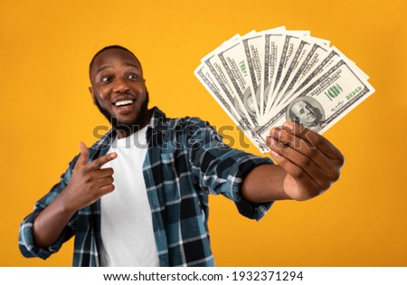 Happy Black Man Holding Money Cash Pointing Finger At Dollar Banknotes In His Hand Standing Over Yellow Studio Background. Financial Success, Profit And Wealth Concept. Selective Focus