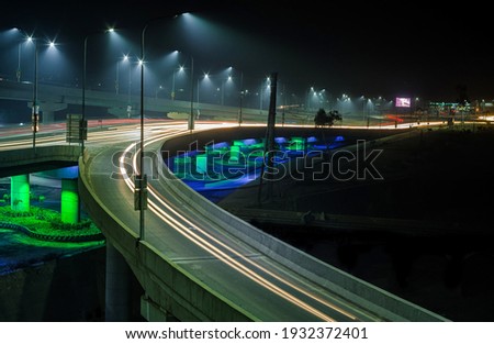 Long exposure photography of flyover in with traffic moving on it and street lights along the road and green neon light below the flyover at night