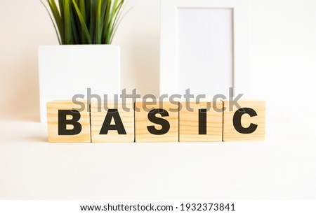 Wooden cubes with letters on a white table. The word is BASIC. White background with photo frame, house plant.