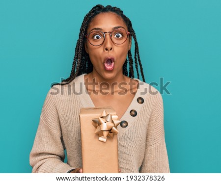 African american woman holding gift scared and amazed with open mouth for surprise, disbelief face 