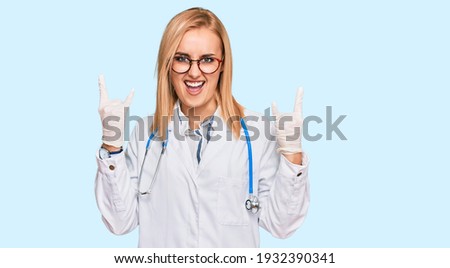 Beautiful caucasian woman wearing doctor uniform and stethoscope shouting with crazy expression doing rock symbol with hands up. music star. heavy concept. 
