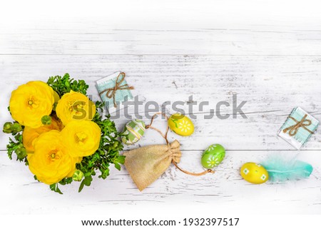 Gorgeous yellow ranunculus buttercups, greeting boxs, Easter painted eggs, feather, canvas bag of gifts on a white wooden background. Easter card celebration.