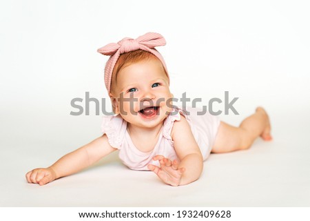 Portrait of adorable red-haired baby lying on belly, white background, wearing pink body and headband Royalty-Free Stock Photo #1932409628