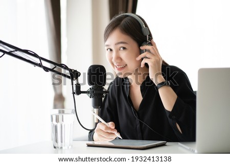 Happy Asian woman setting up a living room in her house for podcast studio, woman arranging a podcast and online radio station at home. Professional young podcaster speaking through a microphone.
