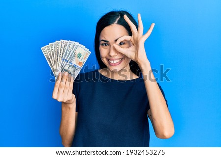 Beautiful young woman holding dollars doing ok gesture with hand smiling, eye looking through fingers with happy face. 