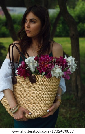 a bouquet of white and red peonies in a bag of a young brunette woman standing in the middle of the park