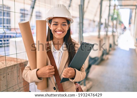 Young latin architect girl smiling happy holding blueprints standing at the city. Royalty-Free Stock Photo #1932476429