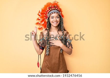 Young beautiful latin girl wearing indian costume smiling swearing with hand on chest and fingers up, making a loyalty promise oath 
