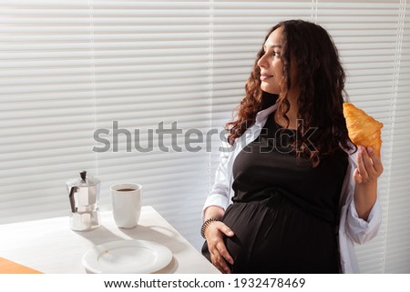 Joyful pregnant young beautiful woman looks through blinds during her morning breakfast with coffee and croissants. Concept of good morning and waiting for meeting with a baby