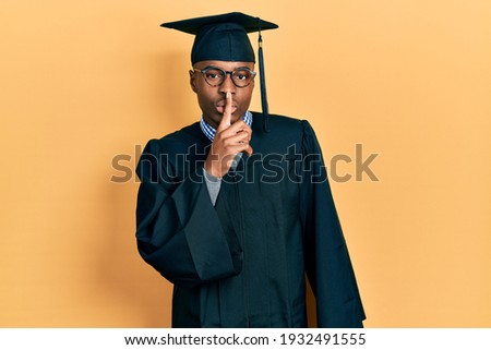 Young african american man wearing graduation cap and ceremony robe asking to be quiet with finger on lips. silence and secret concept. 