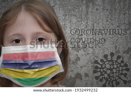 Little girl in medical mask with flag of mauritius stands near the old vintage wall with text coronavirus, covid, and virus picture. Stop virus