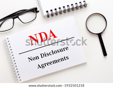 NDA Non Disclosure Agreements is written in a white notebook on a light background near the notebook, black-framed glasses and a magnifying glass.