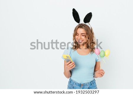 Beautiful happy young woman wearing easter bunny ears holding colored eggs and mobile phone while standing on white studio background with copy space.
