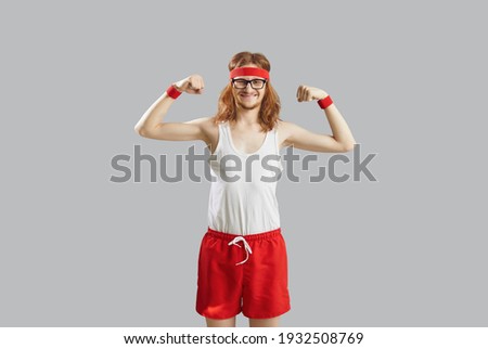 Funny thin skinny man with long hair in eyeglasses, retro headband, white tank top and red shorts showing weak muscles standing isolated on gray background. Sports workout and fitness exercise concept