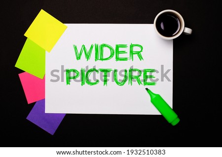 A sheet of paper with the words WIDER PICTURE, a cup of coffee, bright multi-colored stickers for notes and a green marker on a black background. View from above.