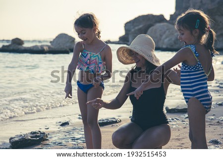 Mom and two little daughters are walking along the seashore in swimsuits, looking at something interesting in the sand. Family vacation at the sea.