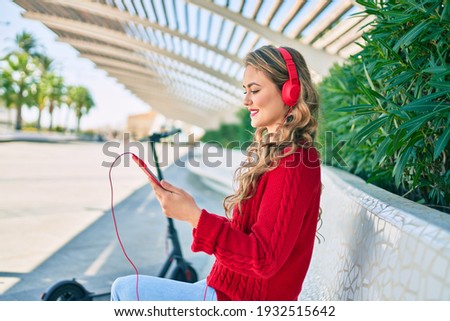 Young blonde girl smiling happy using headphones and touchpad sitting on the bench at the park.