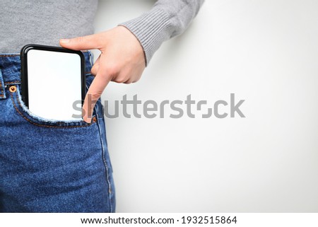 Woman puts mobile phone in jeans pocket on gray light background. Isolation, space for text.  Royalty-Free Stock Photo #1932515864
