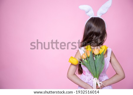 A little girl with easter bunny ears holds a bouquet of tulips in her hands behind her back on a pink studio background.