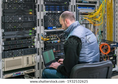A man with a laptop sits in a server room. A technician works near the racks of a modern data center. The system administrator configures the computer hardware. Royalty-Free Stock Photo #1932532625