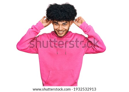 Young african american man with afro hair wearing casual pink sweatshirt smiling pulling ears with fingers, funny gesture. audition problem 