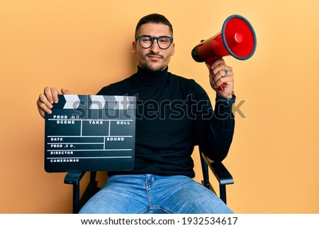 Handsome man with tattoos holding video film clapboard and megaphone skeptic and nervous, frowning upset because of problem. negative person.  Royalty-Free Stock Photo #1932534617