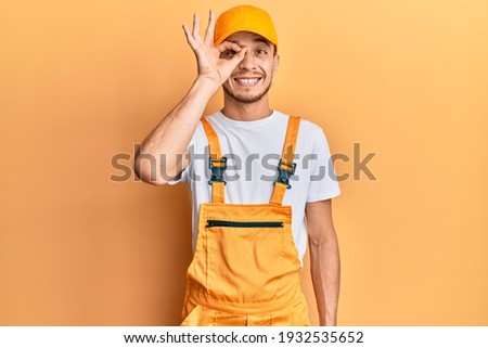 Hispanic young man wearing handyman uniform doing ok gesture with hand smiling, eye looking through fingers with happy face. 