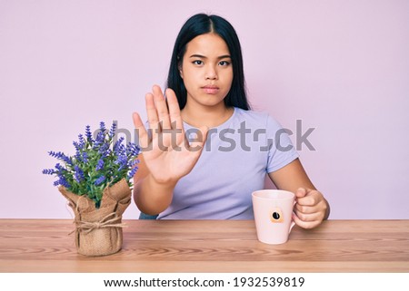 Young beautiful asian girl drinking a cup of infused lavender with open hand doing stop sign with serious and confident expression, defense gesture 