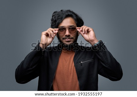 Wearing black jacket and orange shirt hipster person takes off his sunglasses looking at camera and posing in gray background. Royalty-Free Stock Photo #1932555197