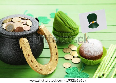 Pot of coins with clover leafs, paper muffin cases, straws, cupcake and flag with picture of St. Patrick's hat on green wooden background
