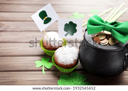 Pot of coins with bow tie, cupcakes and flags with picture of St. Patrick's hat and clover leaf on brown wooden background