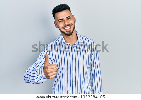Young hispanic man with beard wearing casual striped shirt doing happy thumbs up gesture with hand. approving expression looking at the camera showing success. 