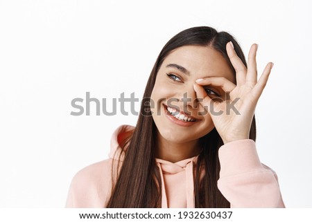 Close up of smiling beautiful woman with natural make up, showing okay sign on eye, looking at upper left corner at logo copy space, seeing interesting banner, white background
