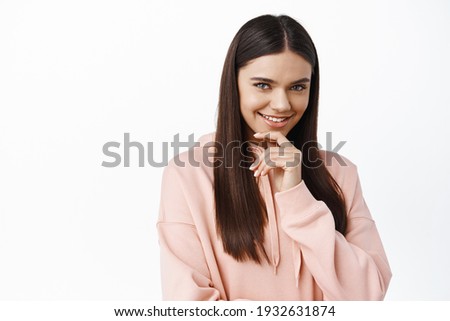 Portrait of pensive brunette woman with long healthy and smooth hair, smiling intrigued, touching chin thoughtful, thinking and staring at camera, white background