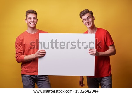 Advertising. Your text here. Couple of handsome young men holding an empty board with copy space together on yellow background.