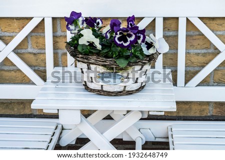 Pansies in a basket on white garden furniture decorate the entrance to the house. Floral home decoration in vintage farmhouse style.