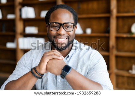 Headshot of friendly and confident African-American guy wearing stylish eyeglasses and smart casual wear, looks at the camera and holds hands in the lock near chin. Employee profile