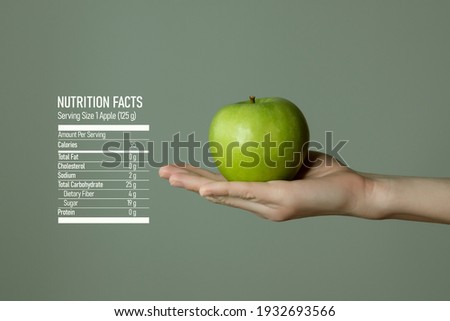 Woman`s hand holding green apple, nutrition facts on grey background. Dietary food and vitamins concept template for product advertising.