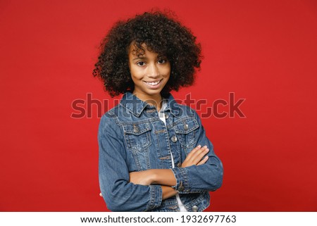 Smiling pretty little african american kid girl 12-13 years old in casual denim jacket holding hands crossed isolated on red color background children studio portrait. Childhood lifestyle concept Royalty-Free Stock Photo #1932697763