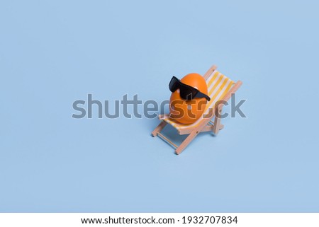 Creative funny composition with Easter egg with sunglasses while sitting on deck chair on pastel blue background. Minimal spring or summer vacation, holiday and travel concept.