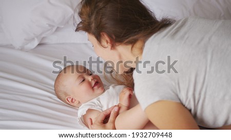 Loving young mommy hugging soothing adorable sweet baby girl lying in bed. Smiling caring mother and cute little infant child girl cuddling in bedroom. Mum and child tender moments.