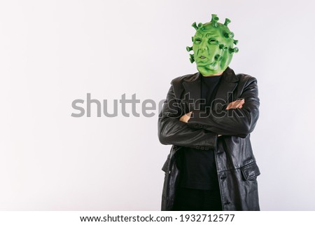 Person disguised as coronavirus with latex mask - covid-19 virus, wearing black leather frock coat and black t-shirt, with crossed arms, on white background. Coronavirus concept