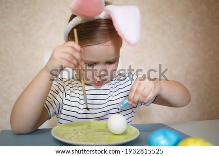 little cute blue-eyed girl in bunny ears paints Easter eggs.