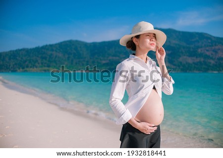 A beautiful asian pregnant women with hat at sandy beach with mountain blue sky and beach as background.
