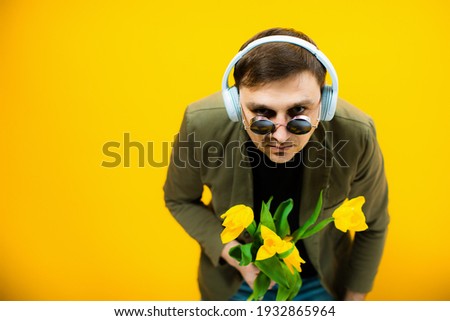 Beautiful macho young man in a jacket with headphones and glasses holding a bouquet of original flowers, withered tulips on a yellow background. Men go home after work and makes caricature emotions