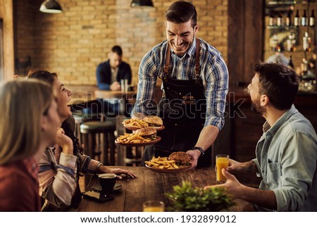 Group of happy friends having fun while waiter is serving them food in a pub. Focus is on waiter.  Royalty-Free Stock Photo #1932899012