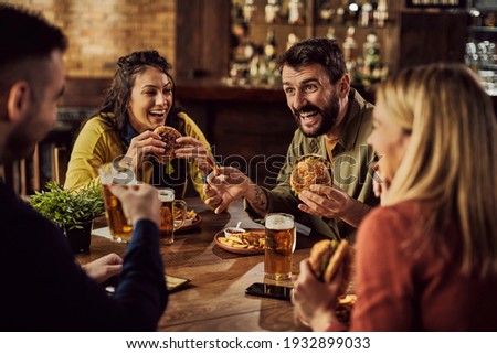 Young happy man having fun with is friends while eating hamburgers and drinking beer in a pub. Royalty-Free Stock Photo #1932899033