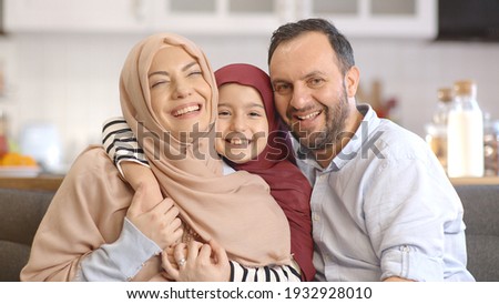 The Muslim little girl wearing a headscarf behind her pregnant mother and father makes a pair of ear sign on their heads and laughs as a family. Happy family portrait. 