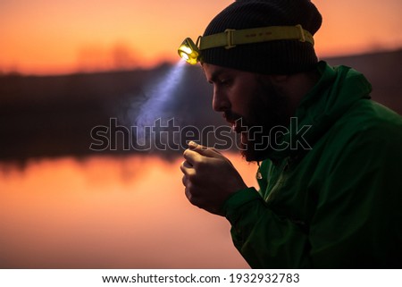 Side view of bearded male traveler in warm clothing and glowing headlamp warming up hands with breath while standing against blurred lake, in nature at sunset time Royalty-Free Stock Photo #1932932783